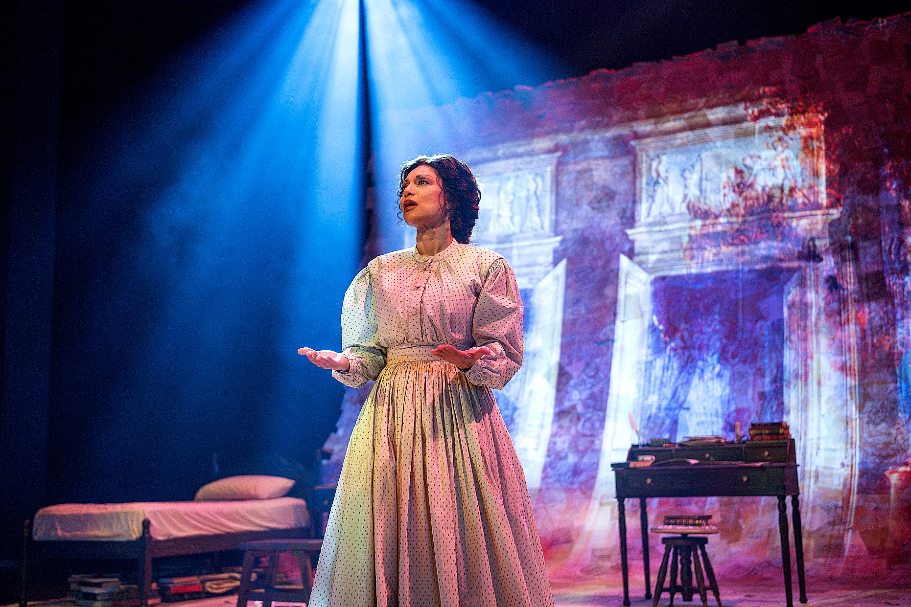 A woman in a cream period dress stands under a vibrant blue spotlight center stage, with her hands turned upwards. To her right is a wooden desk with a small lamp, and to her left is a bed. The background shows a digital projection of historical ledger lines and text.