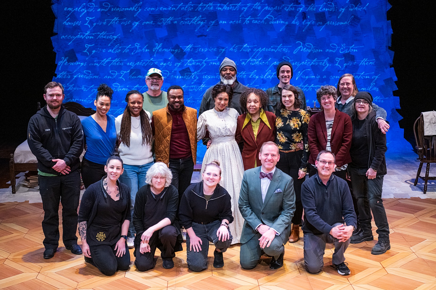 A large group of approximately twenty people poses on a theatre stage. They are arranged in two rows some standing and some kneeling. The background is a bright blue wall projected with elegant cursive handwriting. The individuals are dressed in a mixture of professional attire and theatrical period costumes.