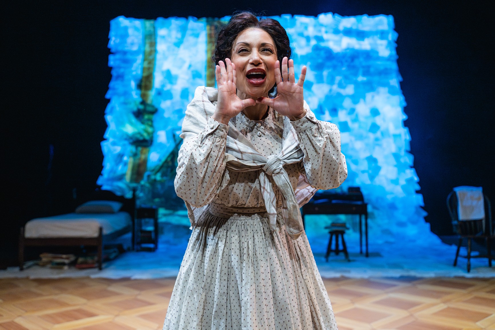 A close-up of a woman on stage wearing a patterned cream dress and a light-colored shawl tied across her chest. She is cupping her hands around her mouth as if calling out. The background is bright, textured blue projection, and the stage floor features a light-colored wood parquet pattern.