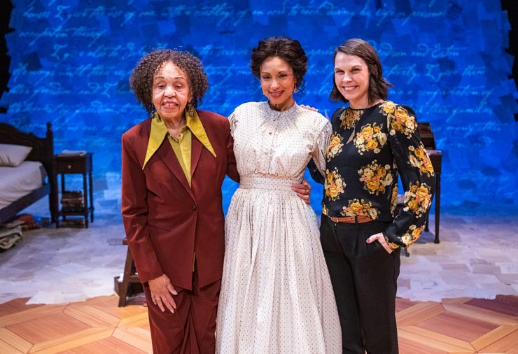 Three women stand together on a stage. The woman on the left wears a maroon blazer and trousers with a gold blouse. The woman in the center wears a cream-colored 19th-century period dress. The woman on the right wears a black floral blouse and black trousers. They are positioned in front of a blue projected backdrop.