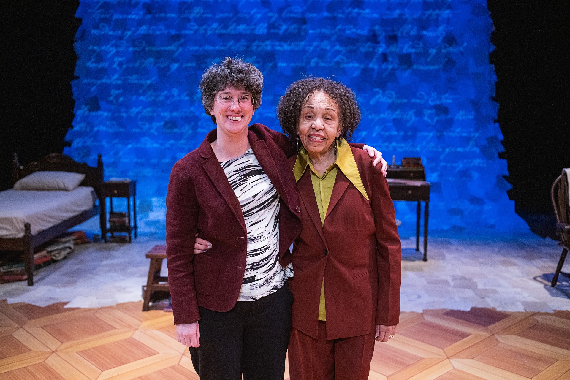 Two women stand close together, smiling, on a theatre stage. The woman on the left wears a maroon blazer over a black and white patterned shirt. The woman on the right wears a maroon suit jacket. The background features a vibrant blue projection of historical context.