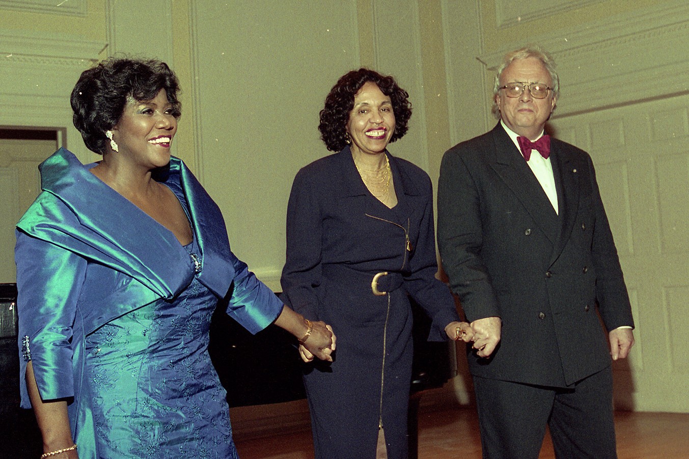 Three individuals stand together. The woman on the left wears an iridescent blue formal gown. The woman in the center wears a dark navy dress. The man on the right wears a black tuxedo with a red bowtie. They are holding hands, smiling towards the audience.