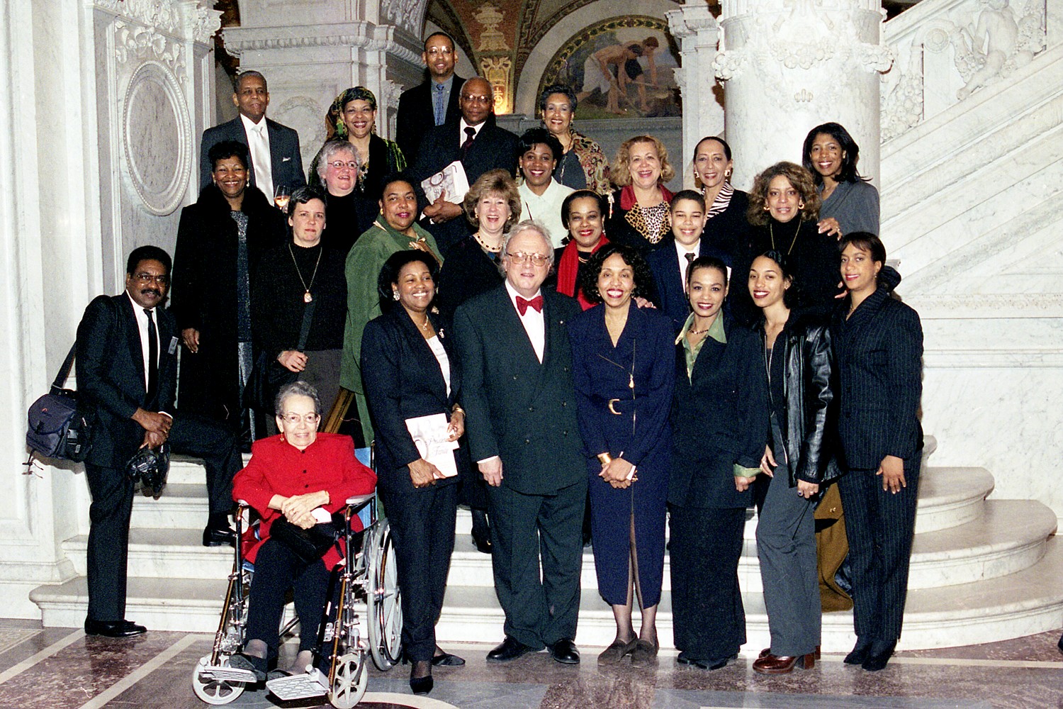 A large group of about twenty people is posed on a grand, ornate marble staircase. The architecture features classical white columns and a decorated archway. The individuals are dressed in a variety of formal and business-casual attire.