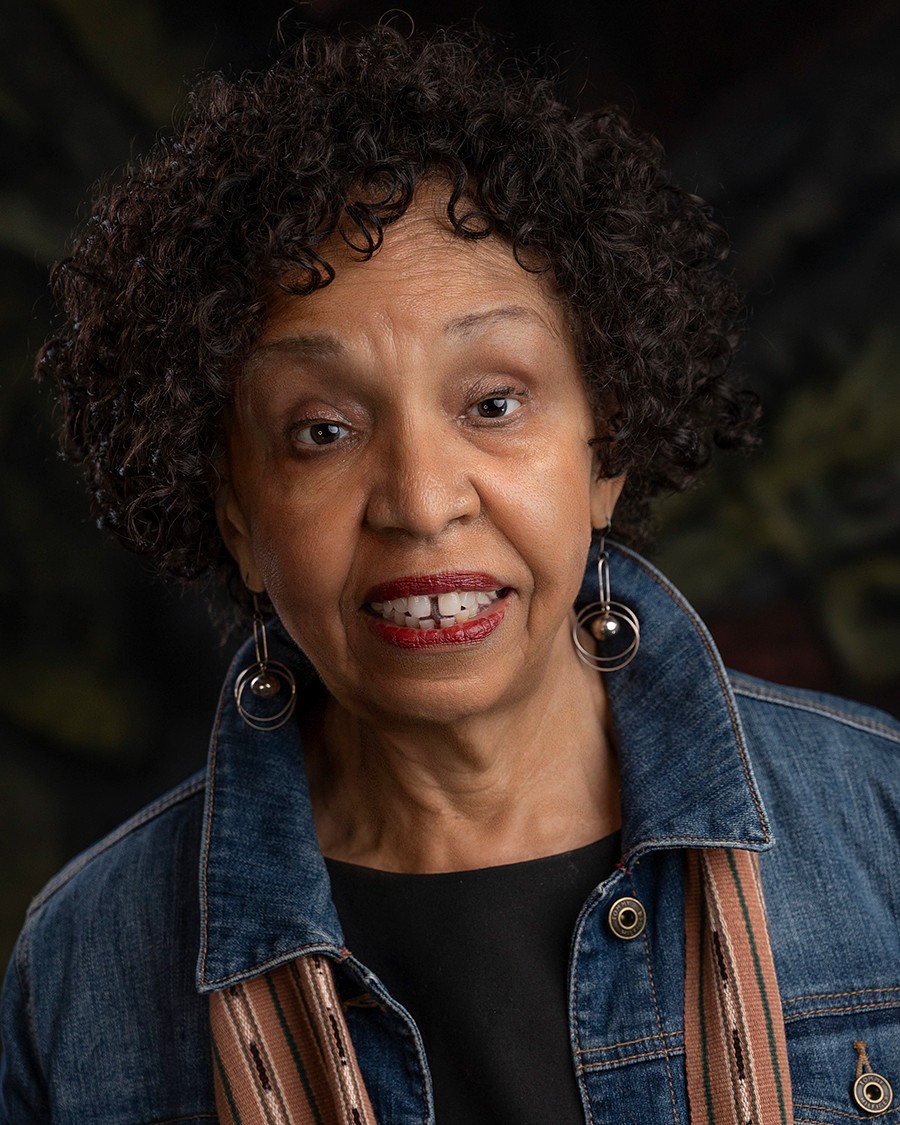Close-up portrait of a woman with short, curly hair wearing a denim jacket, dark top, and dangling earrings, looking directly at the camera against a dark, softly blurred background.