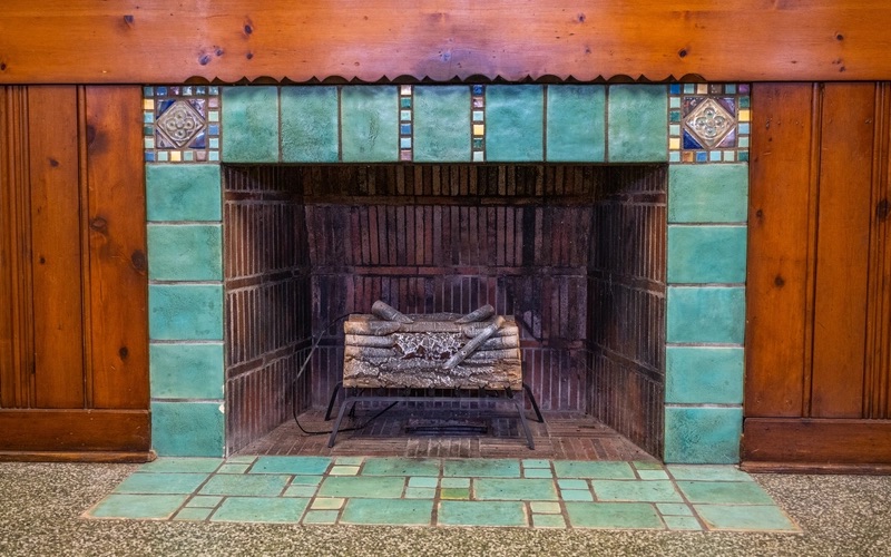 A large fireplace with large green tiles and smaller colorful tiles, is centered in a wooden paneled wall. The inside of the fireplace is lined with thing rectangular brown tiles.