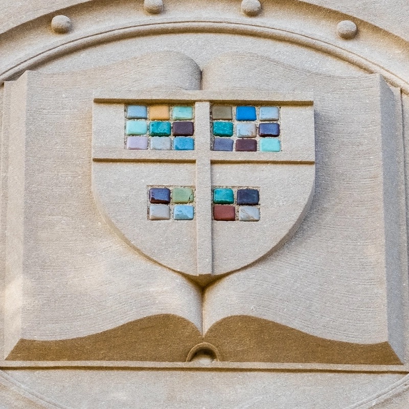 A close up of a stone shield with colorful Pewabic tiles arranged in the center. The shield sits atop an open book, with etchings of lines, depicting writing.