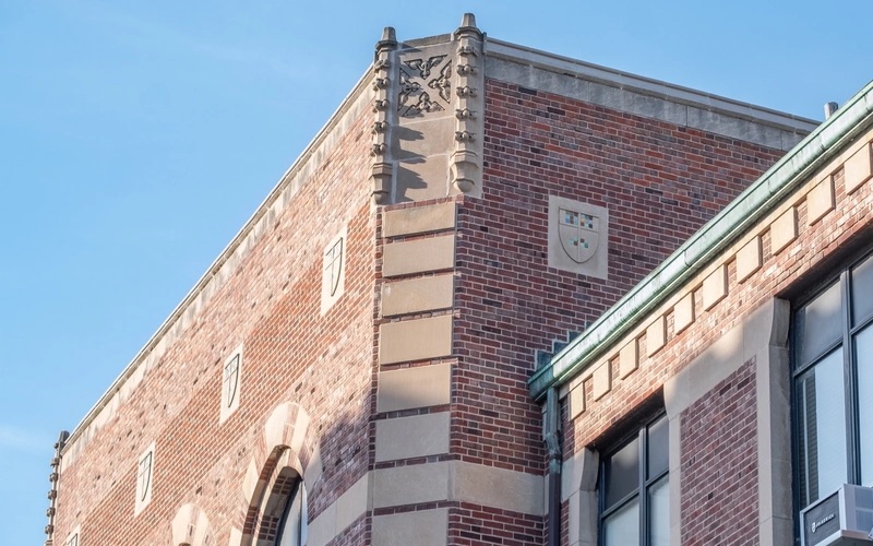 The top of a brick building can be seen against the outline of a clear blue sky. The building is made up of red and brown bricks with designs on the outer corner edge of the highest point. Inlaid into the brick are cement crests with colorful tiles on the inside of the shield-shape.