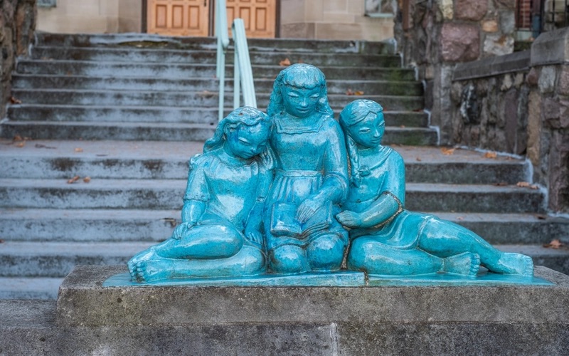 A blue-glazed statue of three children kneeling and leaning against each other. The child in the middle holds an open book in her lap.