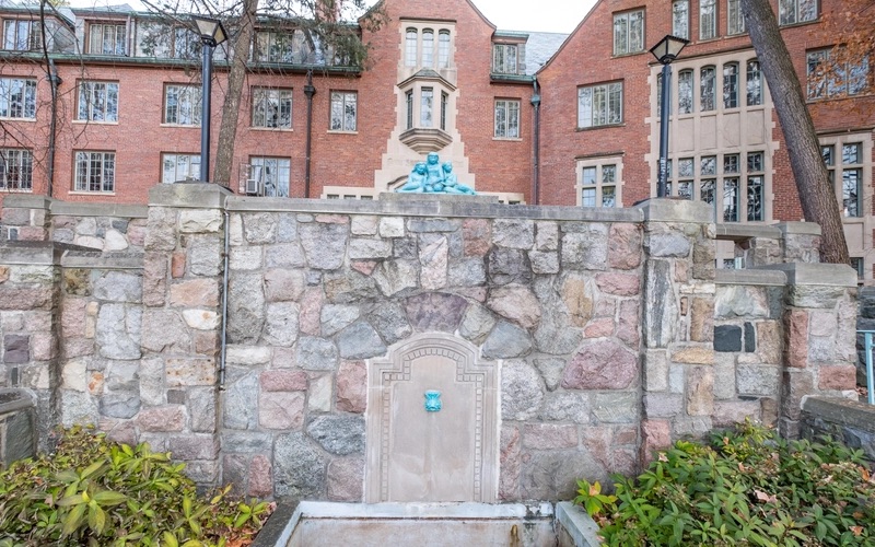A large wall made out of stones, with a blue sculpture of three children sits on top. In the center of the wall is a stele with a blue facet. Behind the wall a large brick building with windows can be seen.