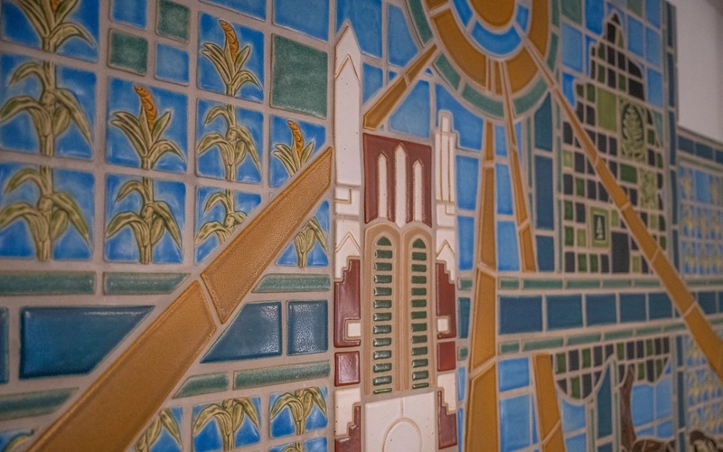 A close-up of a tile mosaic. On some of the tiles are stalks of corn, others make up a green tree. The top of a bell tower is made up of white, brown, and green tiles.