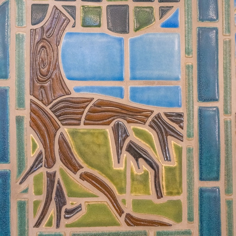A close-up of part of a mosaic. Green, blue, and brown tiles are arranged depicting roots of a tree growing into the earth. The brown tiles that make up the roots have texture and lines that are meant to look like tree bark.
