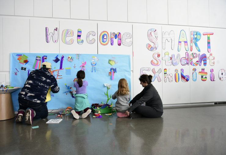 Two adults and two young children sit on the floor and draw on large blue paper adhered to the wall.