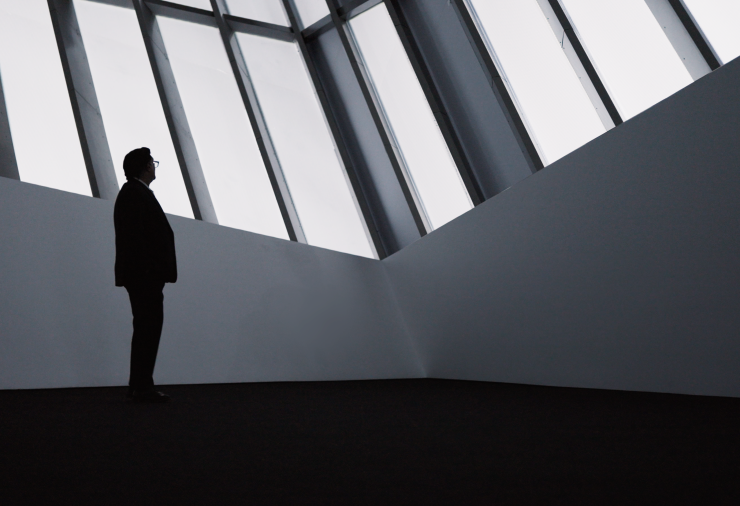 A person stands in a large open gallery space. The room is dark with white light shining against opaque window frames.
