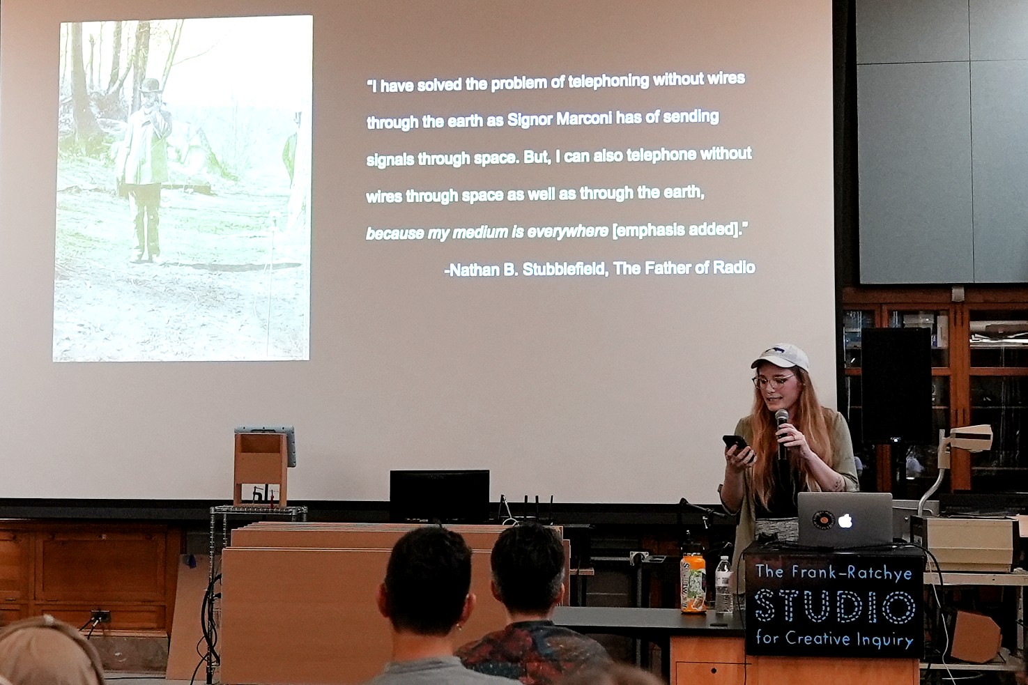A person with long red hair wearing a baseball cap presents a lecture. The projection behind them shows a quote from Nathan B Stubblefield.