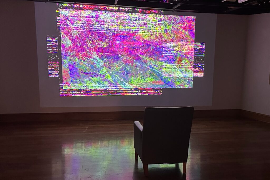 A dimly-lit gallery room with a large-scale video projection on the wall, displaying a colorful abstract image. A single upholstered chair is positioned facing the projection.