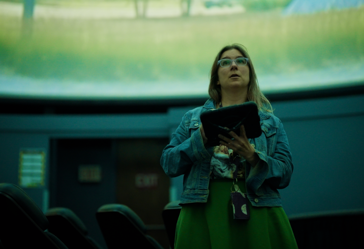 Shannon Schmoll stands in a planetarium holding a tablet, looking up at a projected field.