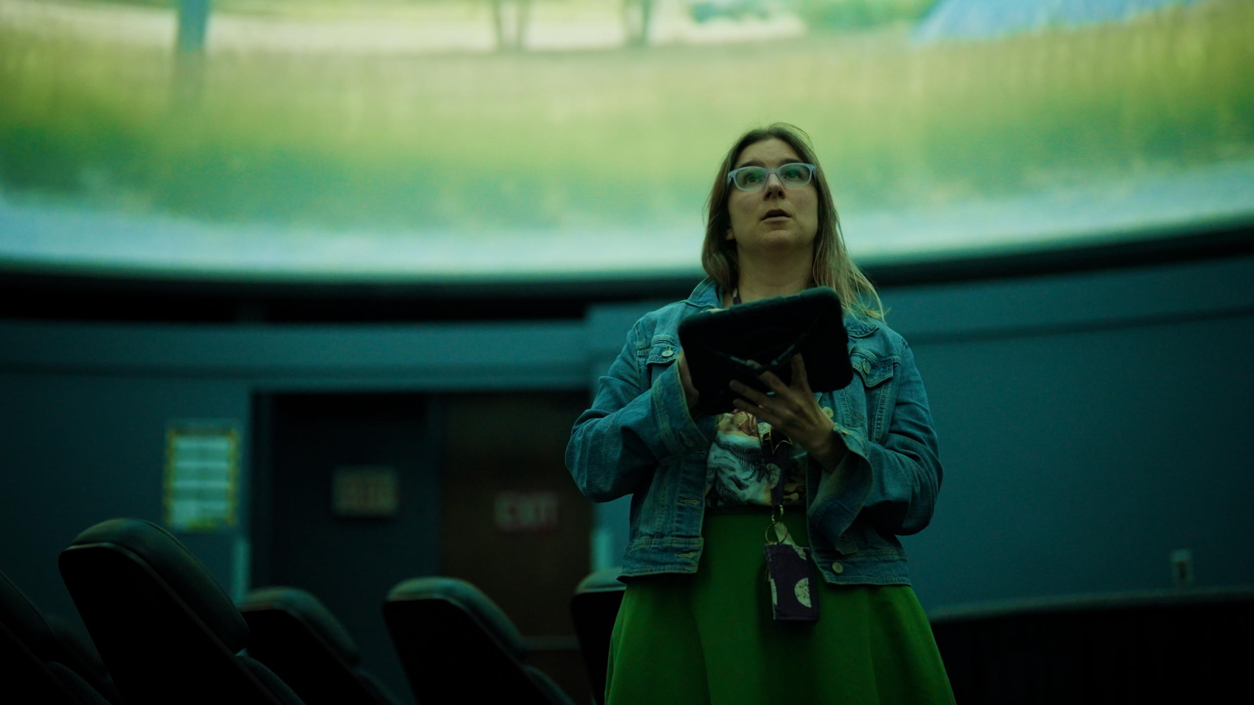 Shannon Schmoll stands in a planetarium holding a tablet, looking up at a projected field.