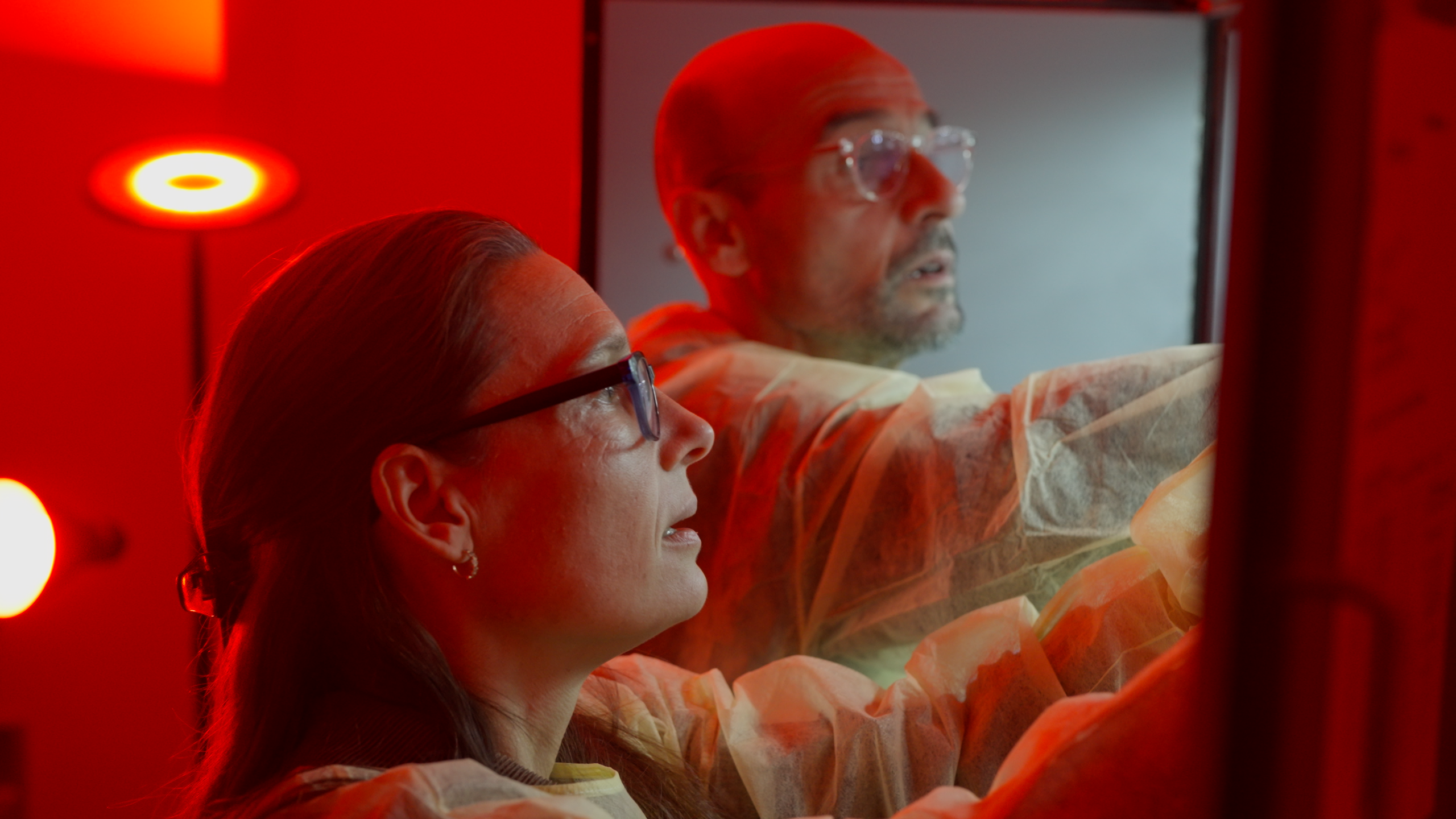 Two people wearing glasses work in a lab with a red light shining behind them.