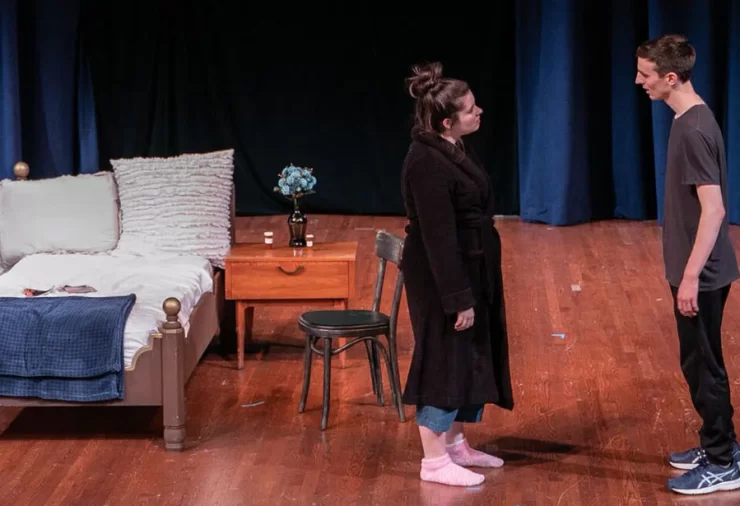 A man in a t-shirt and a woman in a robe stand in front of a theater set with a bed and nightstand.