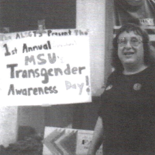 A black-and-white photo of a person with short hair and round glasses, smiling and standing next to a sign that reads