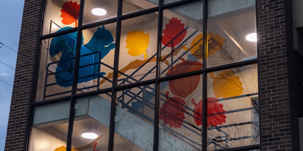 A building with a glass windows exposing the stairwell inside. On the windows are colorful cutouts of a squirrel, acorns, and leaves in a fall color scheme.