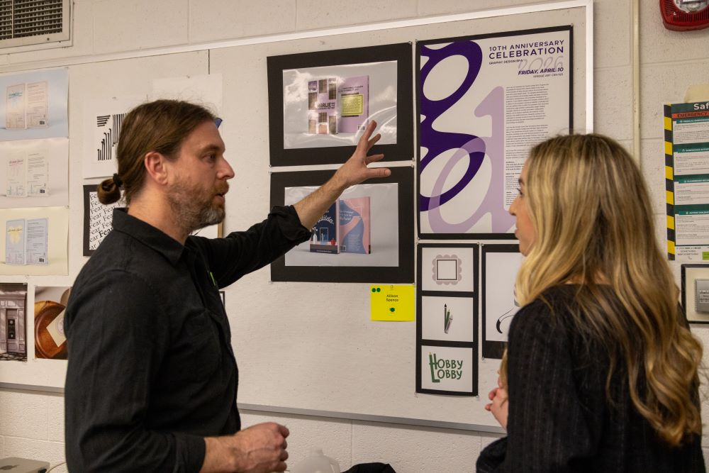 A faculty member and a student, both wearing black shirts, discuss graphic design artwork displayed on a wall.