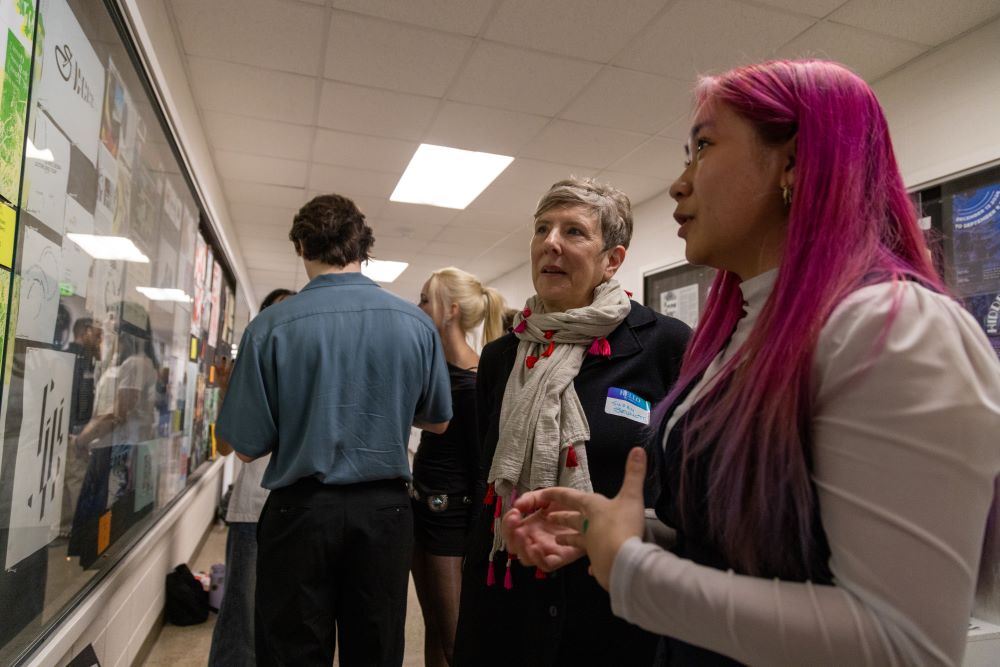 People gather in a hallway for Day of Design. A student with pink hair explains her graphic design project on the wall to an alum wearing a scarf.