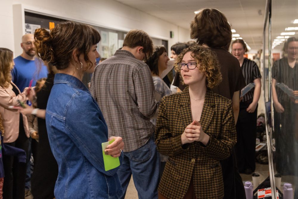 Students and alumni talked about graphic design work displayed on the walls. In the center two women, one in a blue jacket and the other in glasses and a blazer, are in conversation with each other.