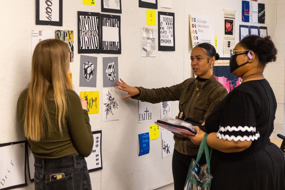 Three people discuss design and typographical work displayed on a wall.