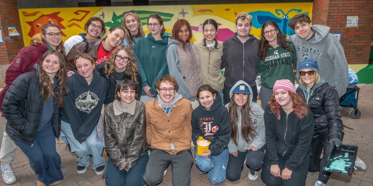 In the background a colorful mural with a yellow background and colorful geometric animals and bugs can be seen. Members of Gamma Epsilon Tau pose as a group in front of the mural.