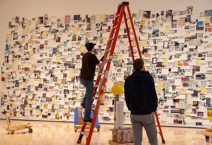 People work on a tall, red ladder in front of a large, collage-style art installation on a wall covered with diverse images and text.