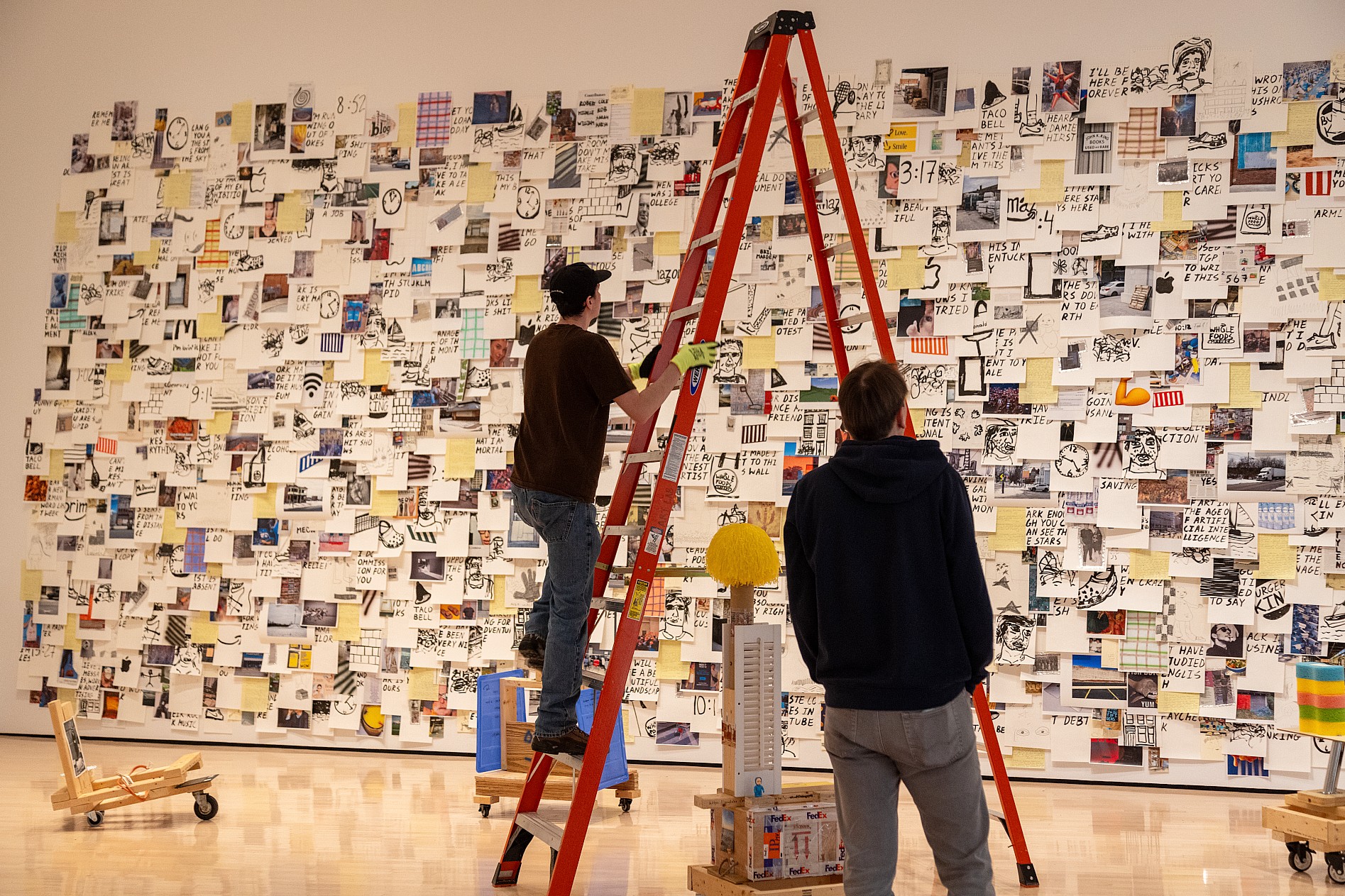 People work on a tall, red ladder in front of a large, collage-style art installation on a wall covered with diverse images and text.