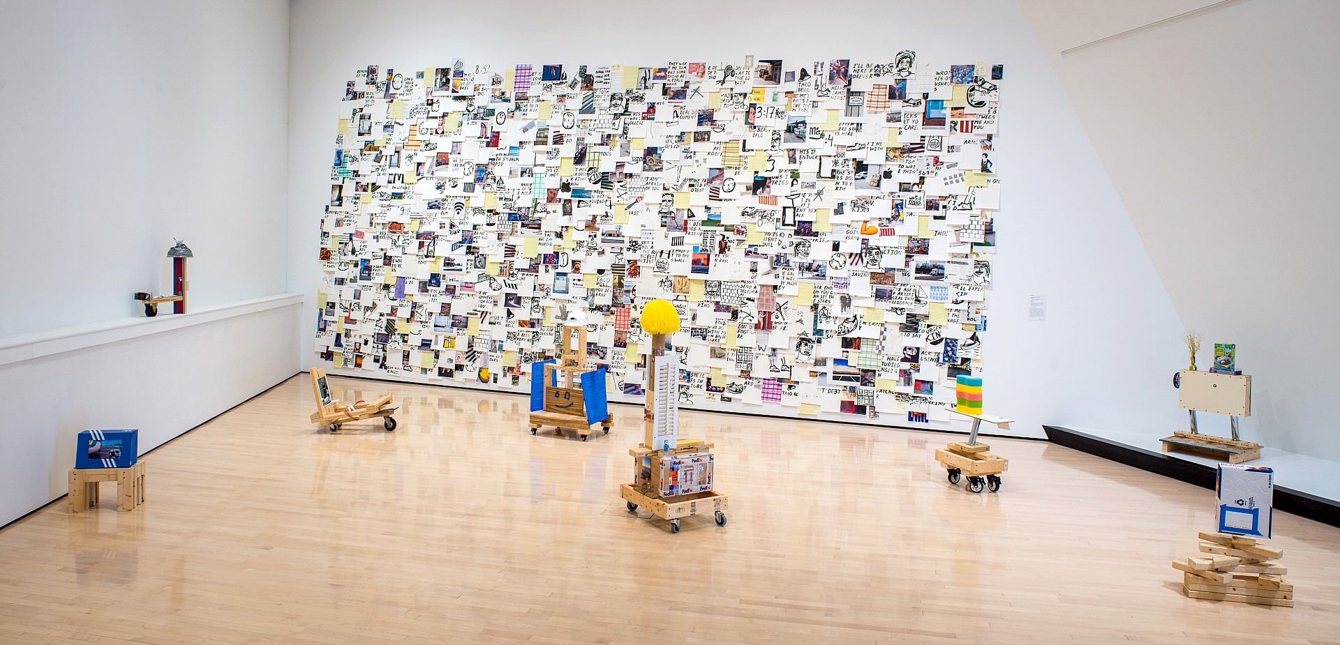 A modern art gallery with a collage wall covered in colorful, eclectic papers. The floor features wooden sculptures on wheels