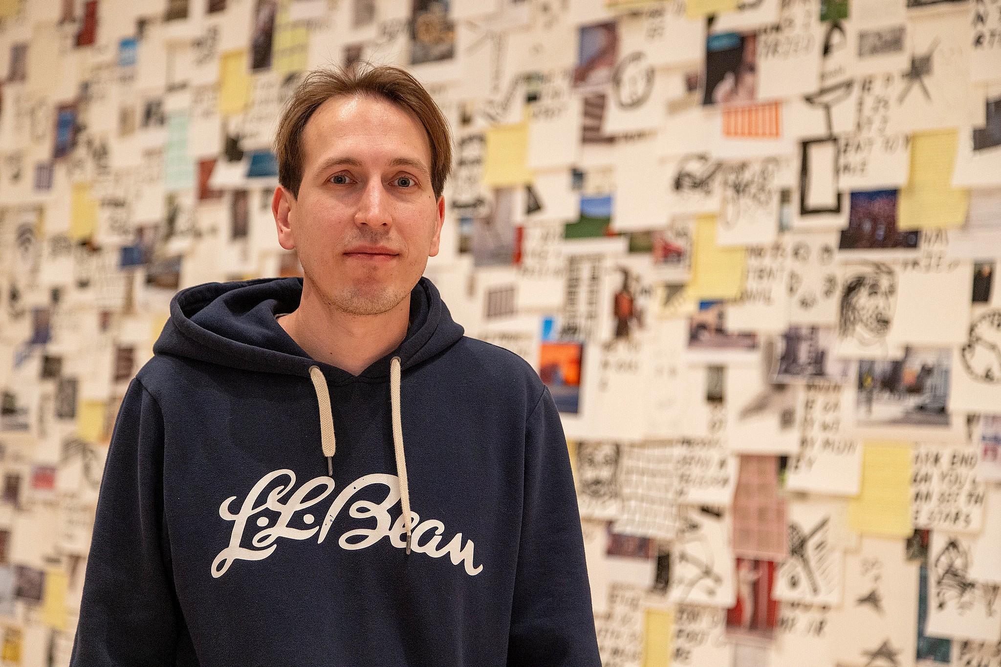A man in a blue L.L. Bean hoodie stands in front of a wall covered with assorted notes and photos, conveying a creative and thoughtful atmosphere.