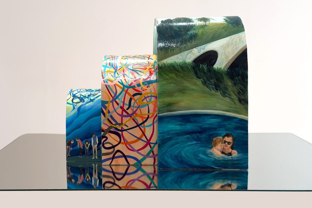 A series of three differently sized, painted sculptures on a reflective surface. Each sculpture features unique artwork: a bridge with water, vibrant swirls, and small figures.