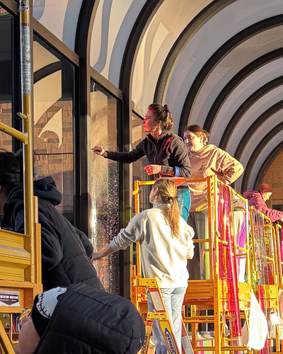 A group of students and faculty stand on scaffolding to install translucent film along windows in a glass arched walkway.