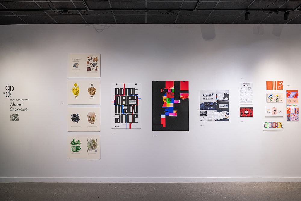 A gallery wall in an exhibition space displays graphic design works, including abstract prints, typography, and colorful poster art. On the wall, the title of the exhibition is: 