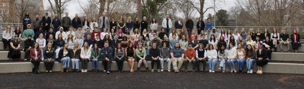 A group of more than 100 students, faculty, and alumni sit outside on risers at the 2026 Day of Design event held at Michigan State University.