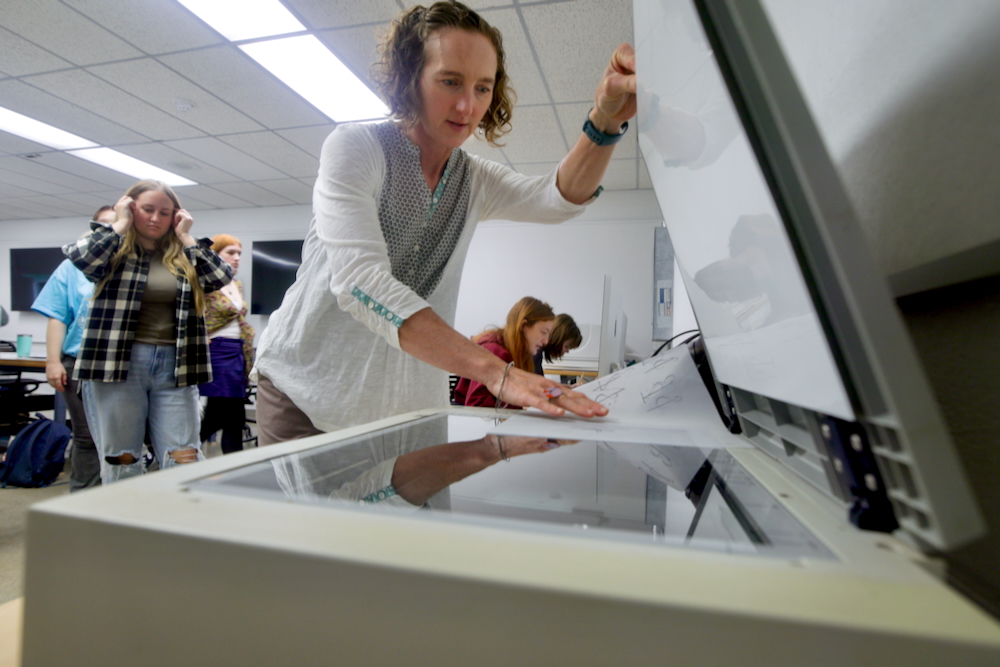 A faculty member in a white shirt places a document in a large scanner in a classroom setting. Several students are in the background watching and ready to participate.