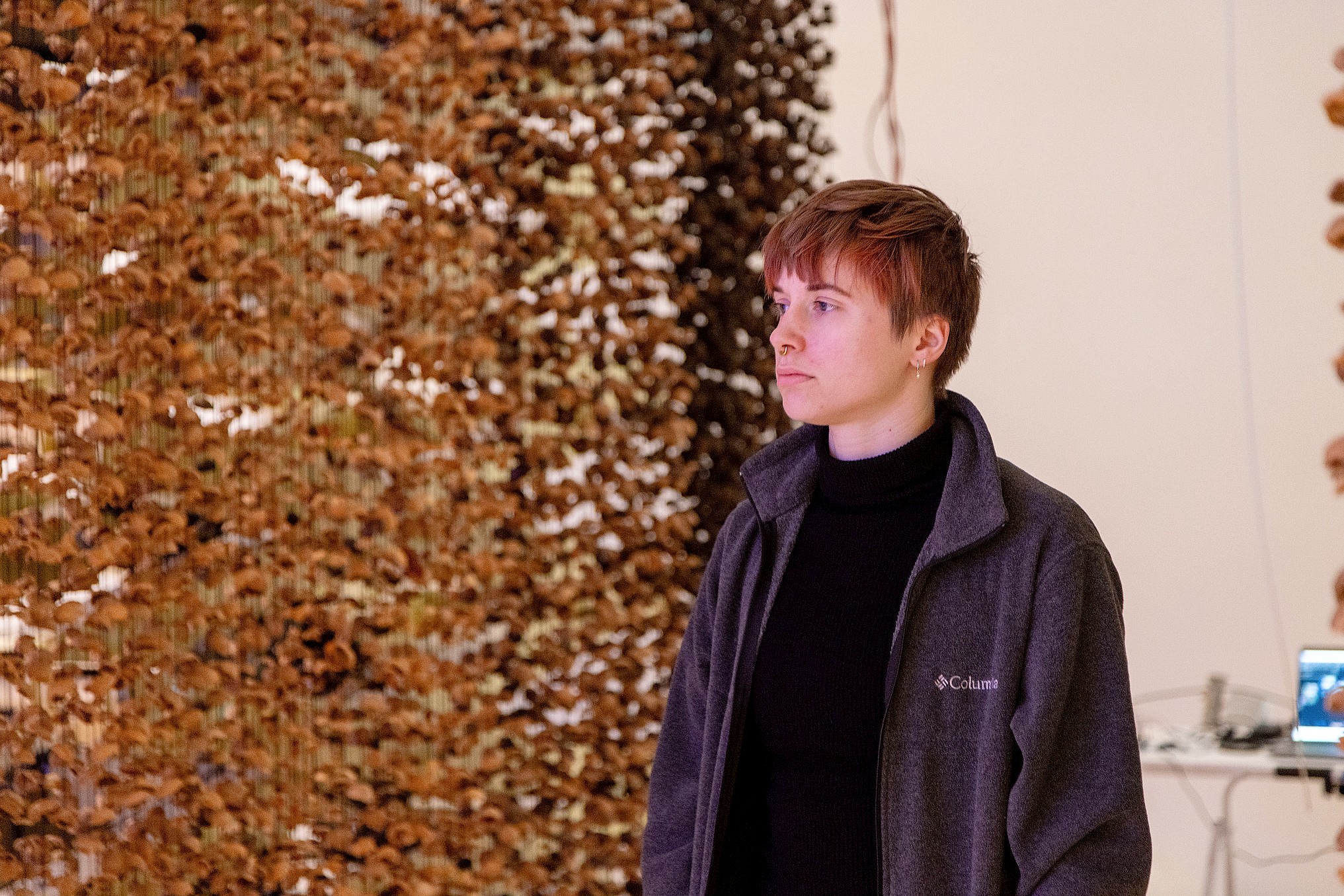 A person with short hair and a gray jacket stands indoors, gazing thoughtfully. Behind them is a large curtain of hanging walnut shells.