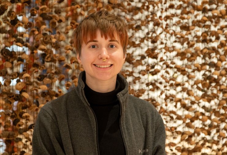 A person with short hair and a septum ring smiles warmly, wearing a dark jacket. Behind them, a textured background of walnut shells creates an earthly atmosphere.