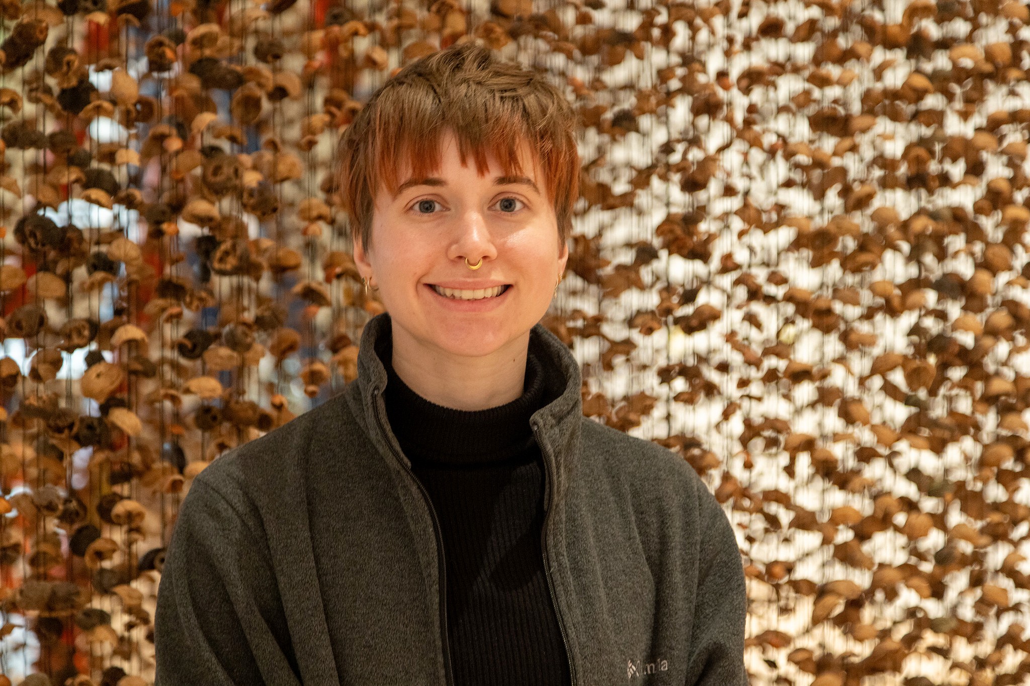 A person with short hair and a septum ring smiles warmly, wearing a dark jacket. Behind them, a textured background of walnut shells creates an earthly atmosphere.