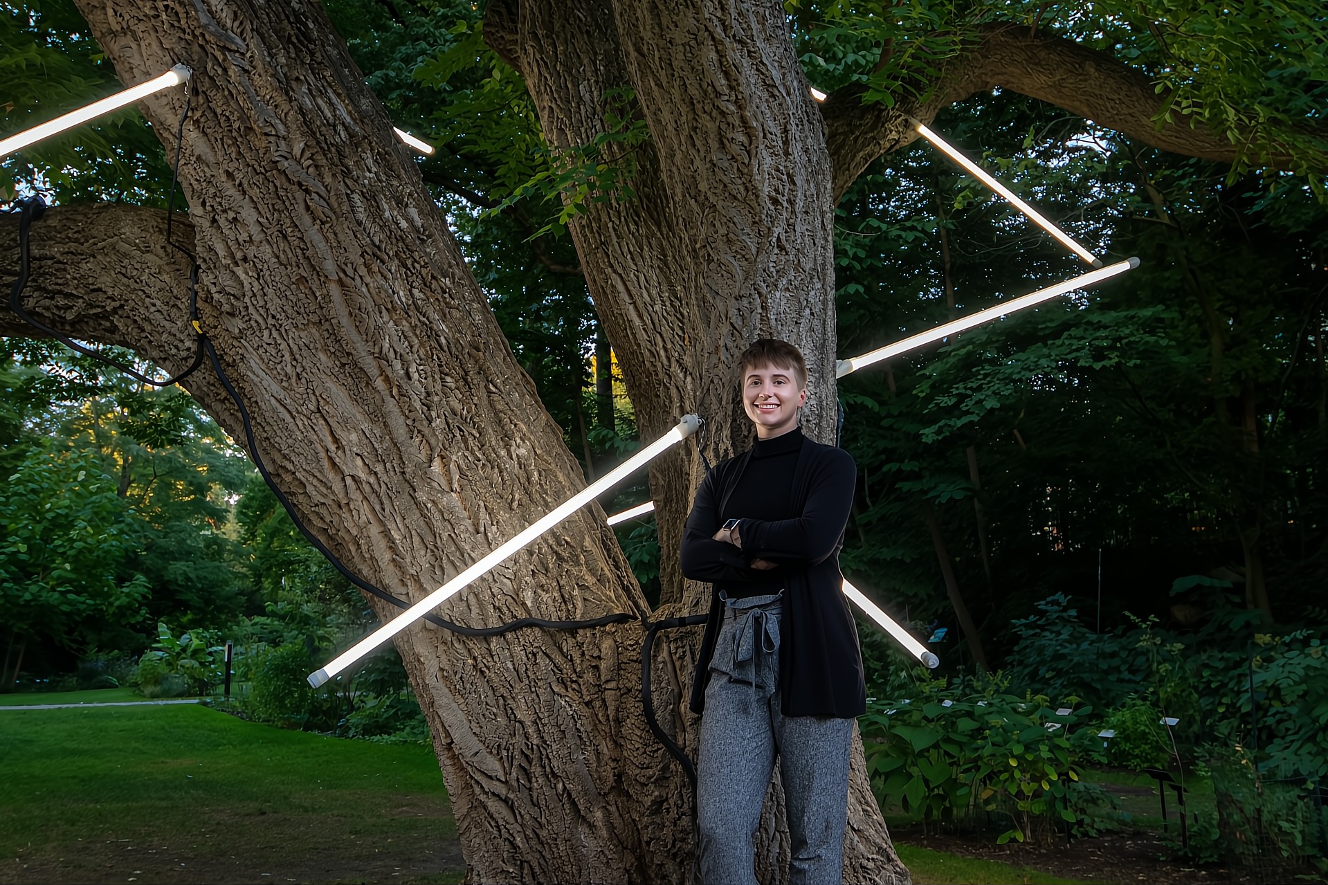 A person wearing a black top and gray pants, standing confidently with crossed arms in front of a tree. Bright light rods are artistically positioned to appear as if they are piercing the tree.