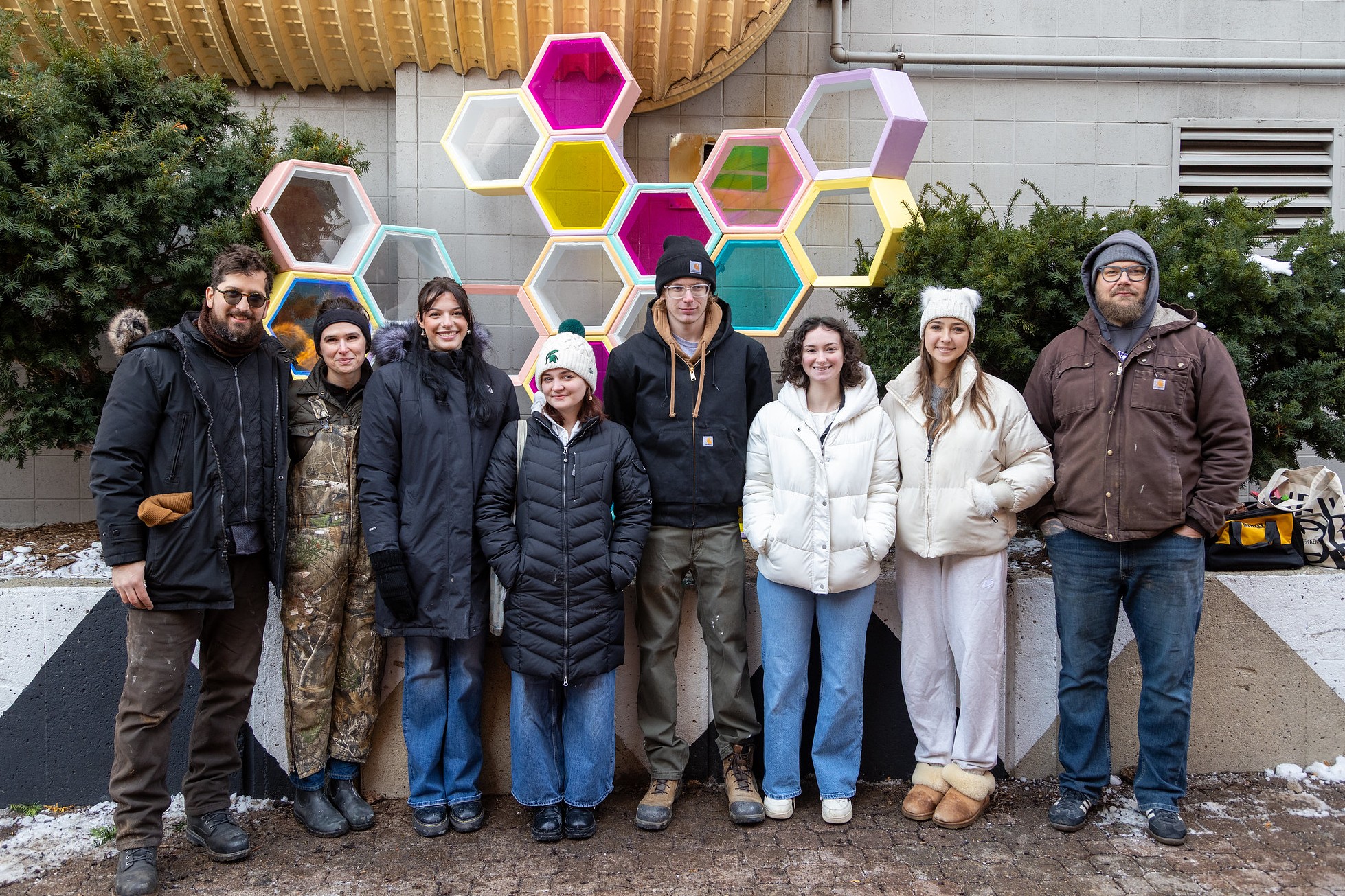A group of eight people stand in winter clothing before a colorful hexagonal art installation. They appear cheerful, with green bushes and a building facade behind them.