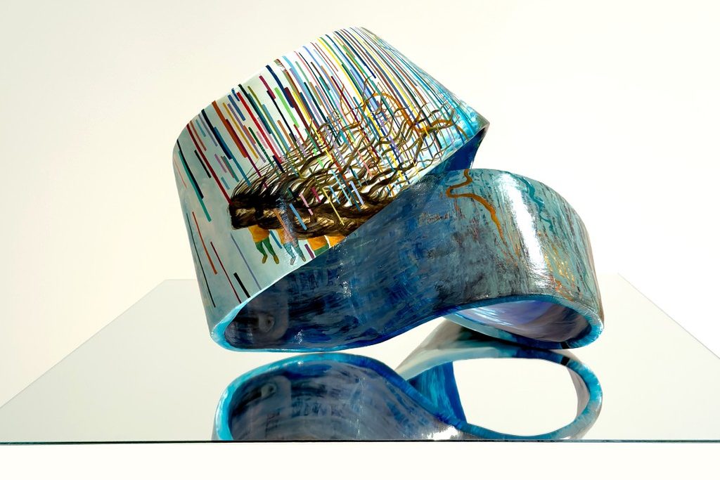 A twisting blue glass sculpture with multicolored vertical lines and metallic accents stands on a reflective surface, creating an elegant, dynamic visual.