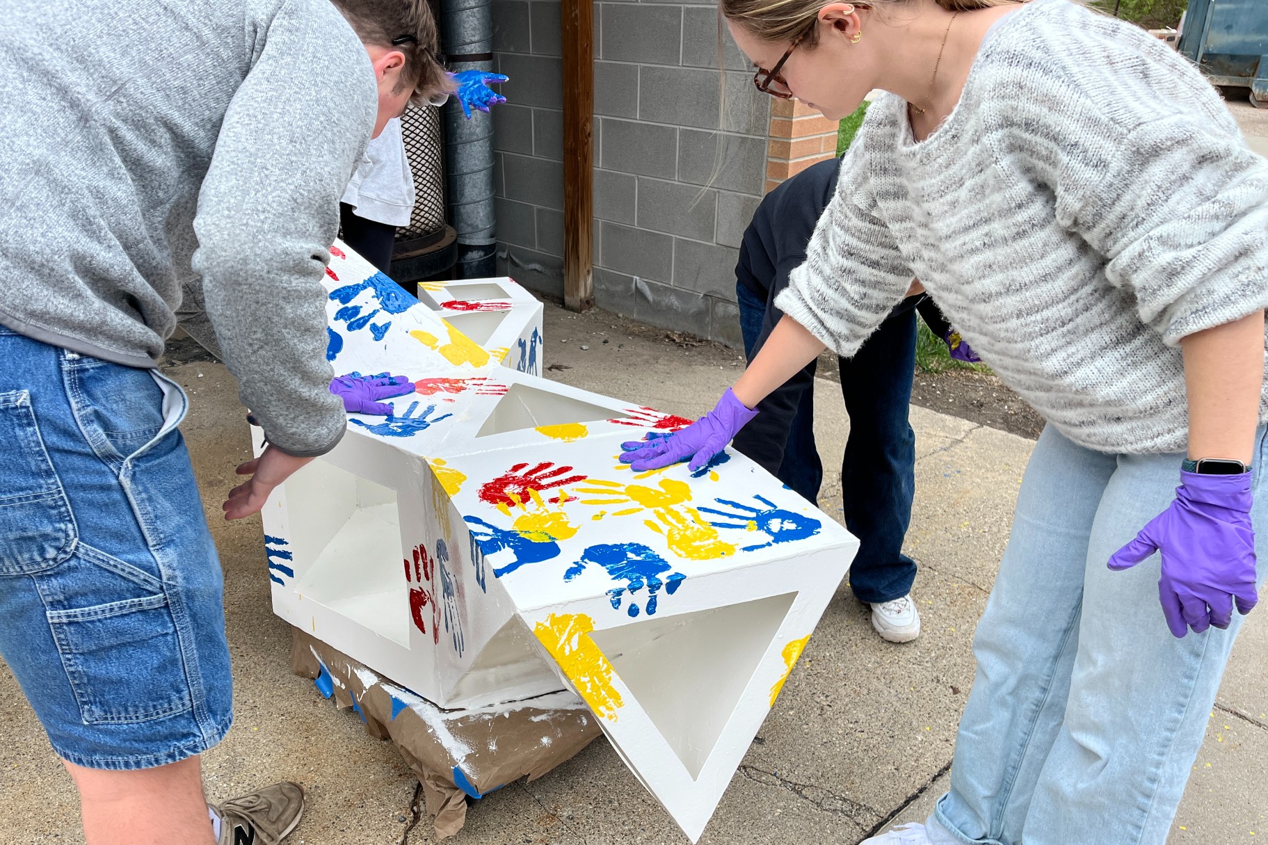 People wearing casual clothing and purple gloves are decorating wooden blocks with colorful handprints. the setting is an outdoor area with a concrete wall.