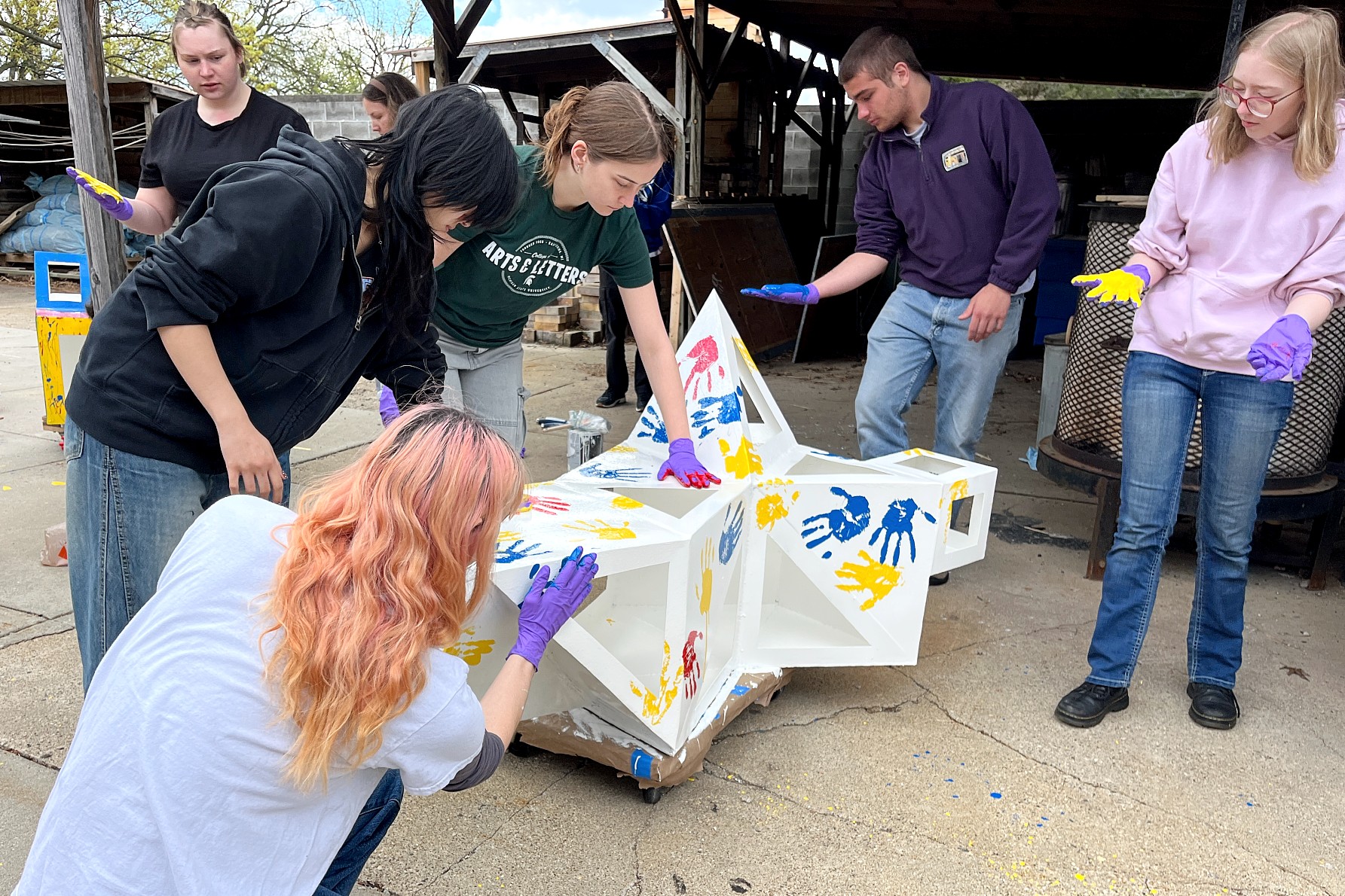 A group of people painting handprints in colorful paints on a white, sculptural object outdoors. They wear gloves and appear focused and collaborative.