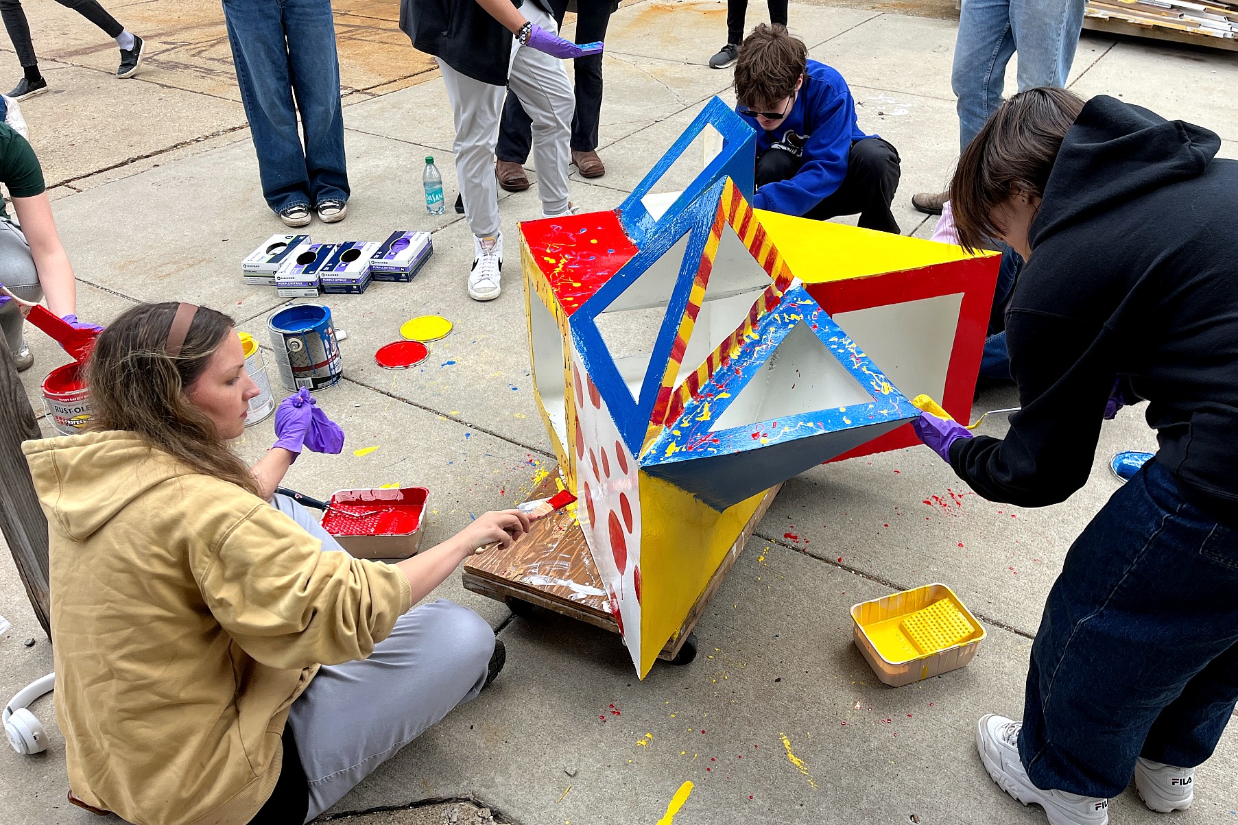 A group of people paint a geometric sculpture outdoors with red, yellow, and blue paint. The scene is lively and collaborative.