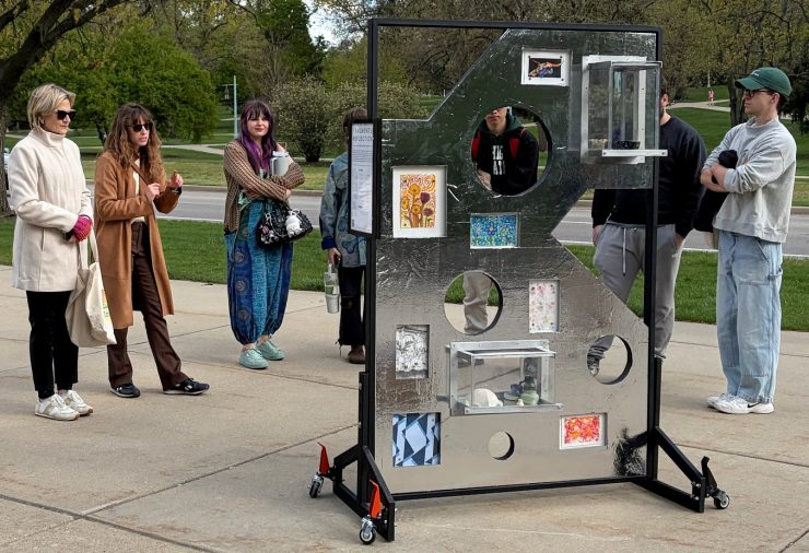 A group of people stands outdoors around a metal art display with various pictures and objects placed in circular cutouts. The mood is curious and engaging.