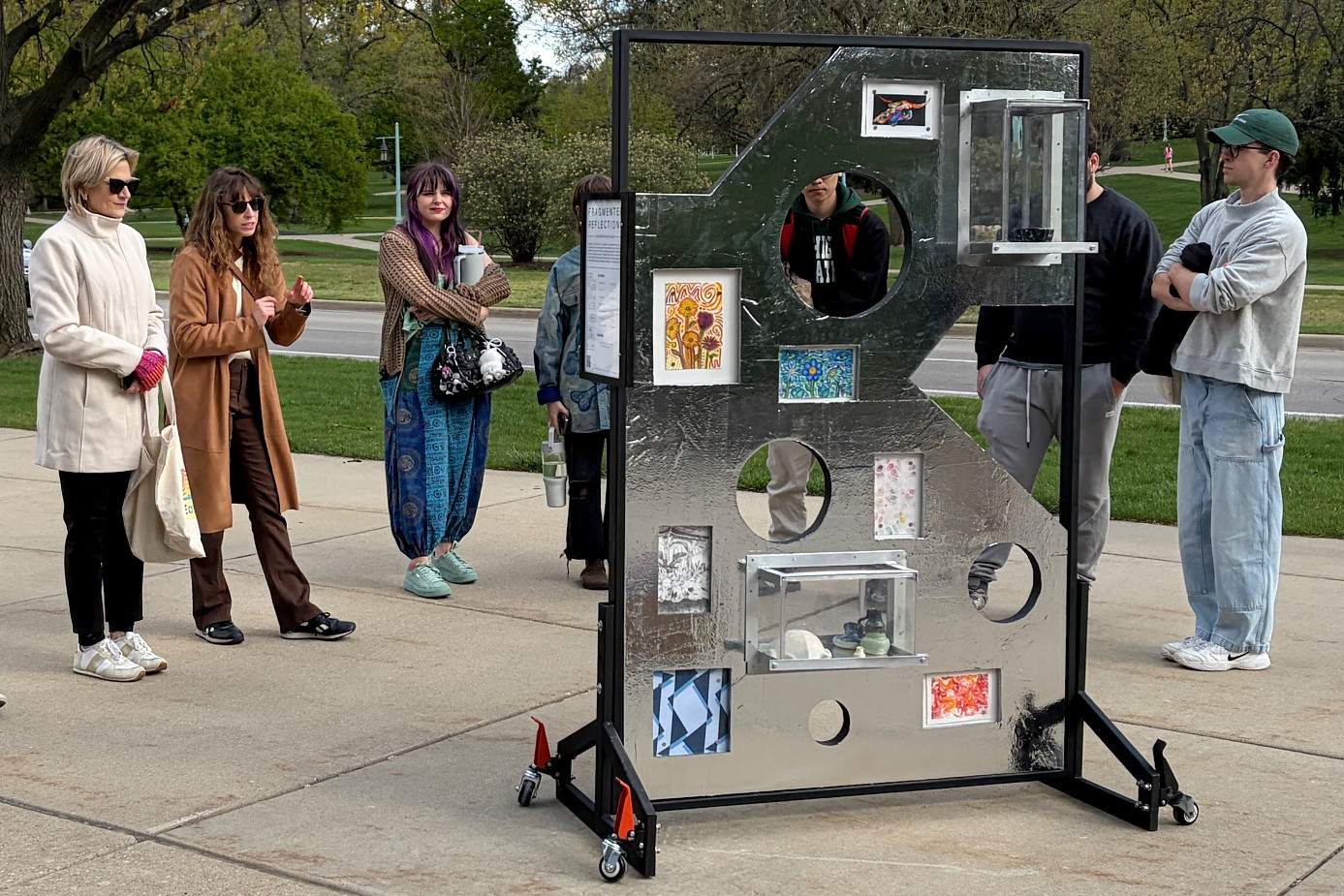 A group of people stands outdoors around a metal art display with various pictures and objects placed in circular cutouts. The mood is curious and engaging.